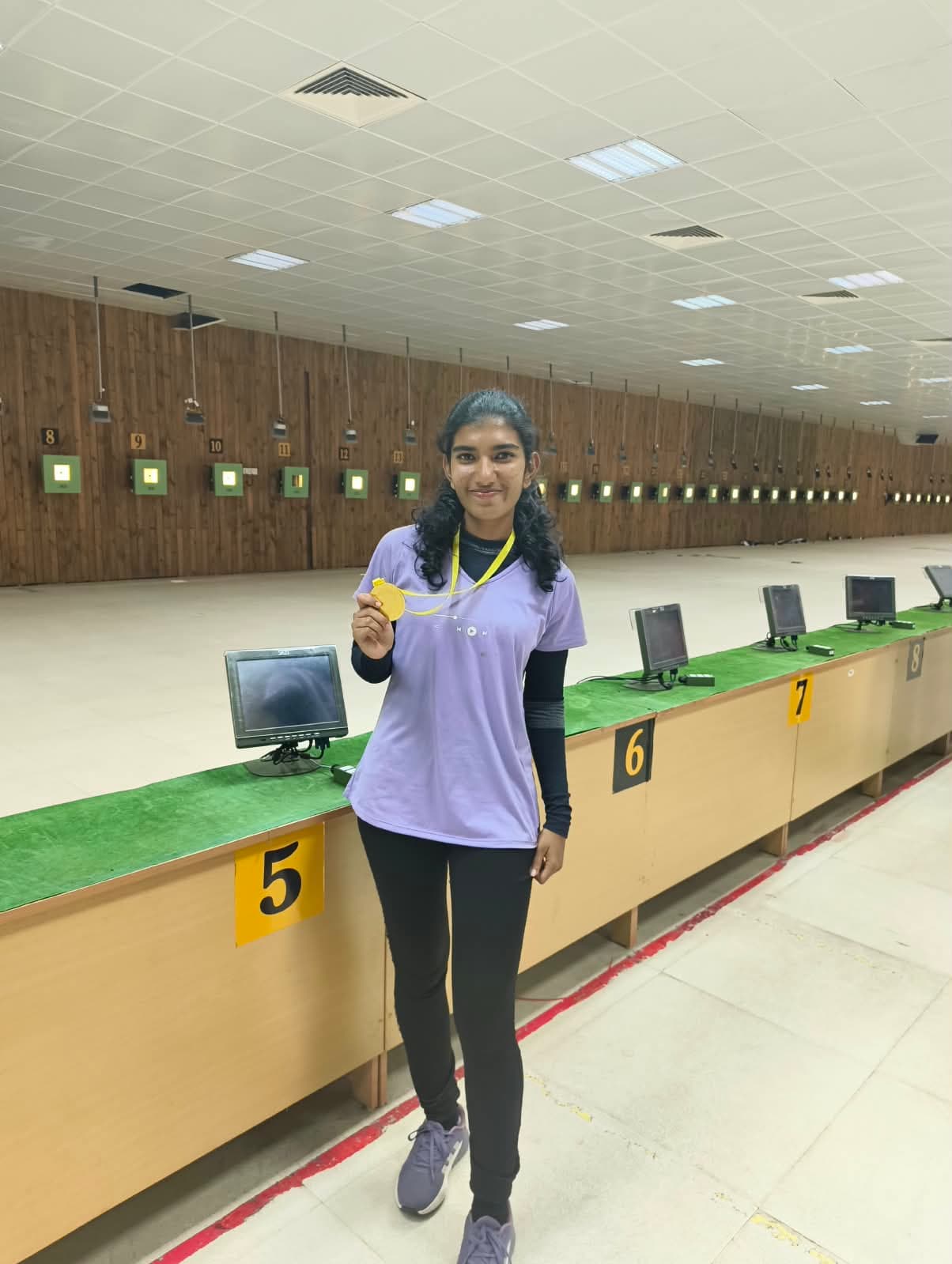 Brahmitha G holding up a gold medal beside the firing points of an indoor range