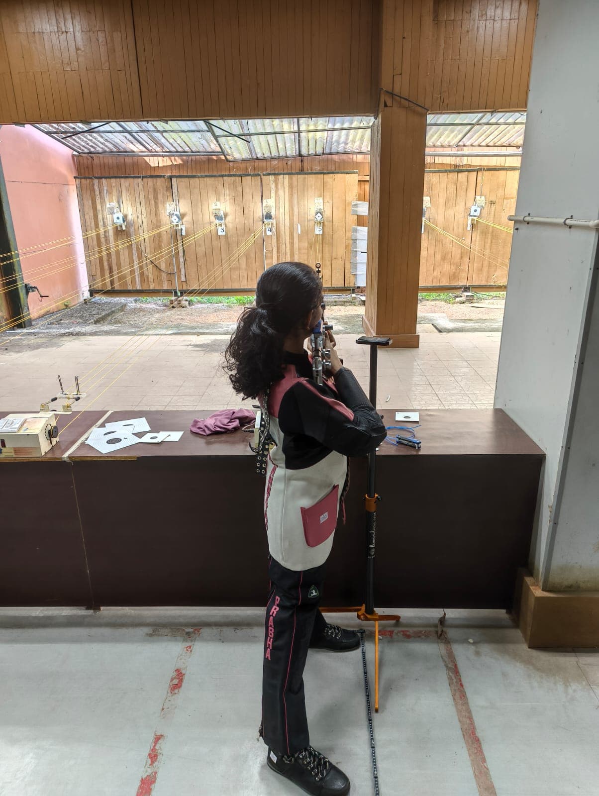 Brahmitha G seen from behind, aiming at paper targets on a wood-panelled practice range